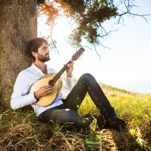Alon sitzt auf einer Wiese, an einen Baum gelehnt und spielt Gitarre