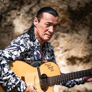 Lulo sitzt vor einem Felsen und hält seine Gitarre in der Hand