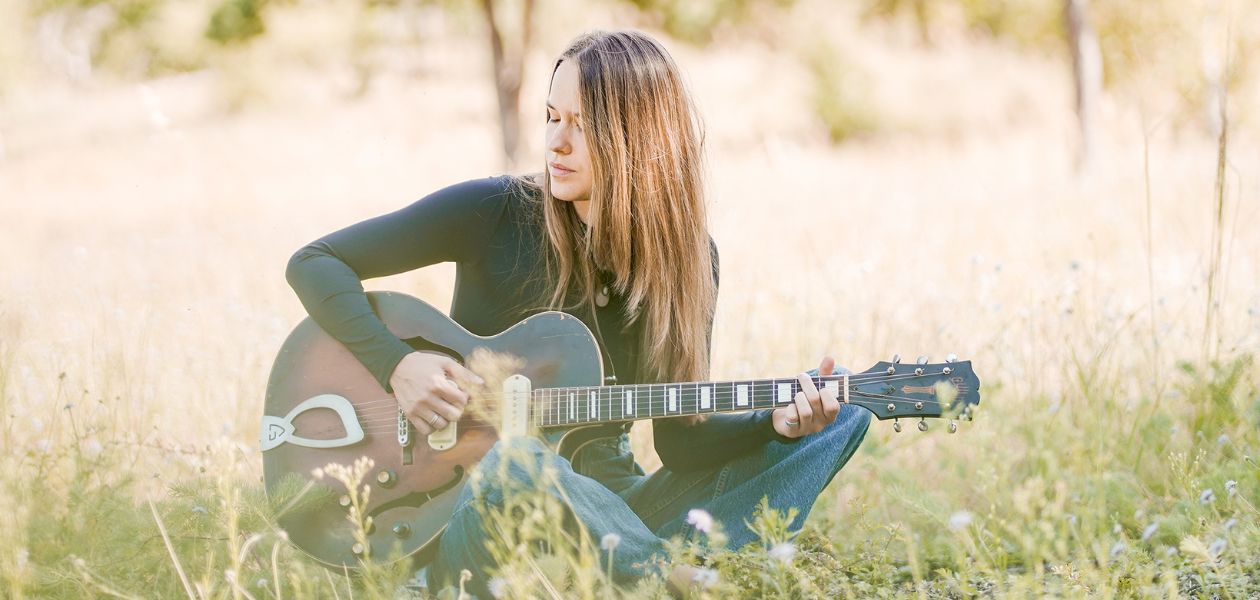 Musikerin Caro Trischler sitzt mit ihrer Gitarre im Gras. Sie hat lange braune Haare und schaut verträumt zur Seite.