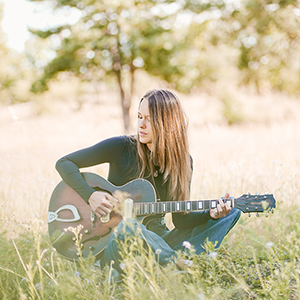 Musikerin Caro Trischler sitzt mit ihrer Gitarre im Gras. Sie hat lange braune Haare und schaut verträumt zur Seite.