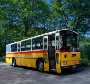 Bild eines historischen gelben Postbusses. Er steht von zwei grünen Bäumen. Der Hintergrund ist KI-generiert.