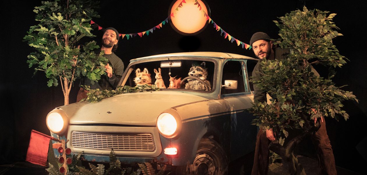 Drei Tiere sitzen in einem alten Auto, zwei Männer schauen rechts und links aus dem Fenster. Über dem Auto scheint der Mond.