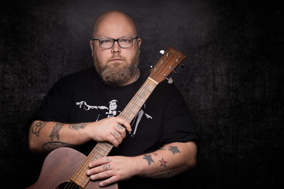 Andreas Kümmert mit Brille und einer Akustikgitarre vor dunklem Hintergrund.