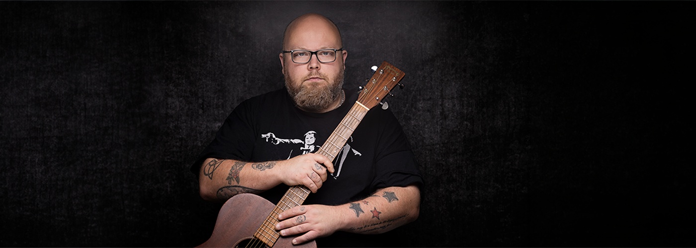 Andreas Kümmert mit Brille und Akustikgitarre vor dunklem Hintergrund.